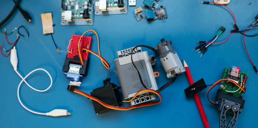 Top-down view of an electronics work surface covered with components used for prototyping and robotics. On a blue mat are several microcontroller boards, small sensor modules, jumper wires, and connectors. There are breadboards with attached wires, a servo motor, and LEGO-style robotic motors and structural parts. We can also see batteries and battery clips, alligator clips, resistors, and small breakout boards arranged loosely rather than assembled into a single circuit. A pencil and various cables are scattered around.