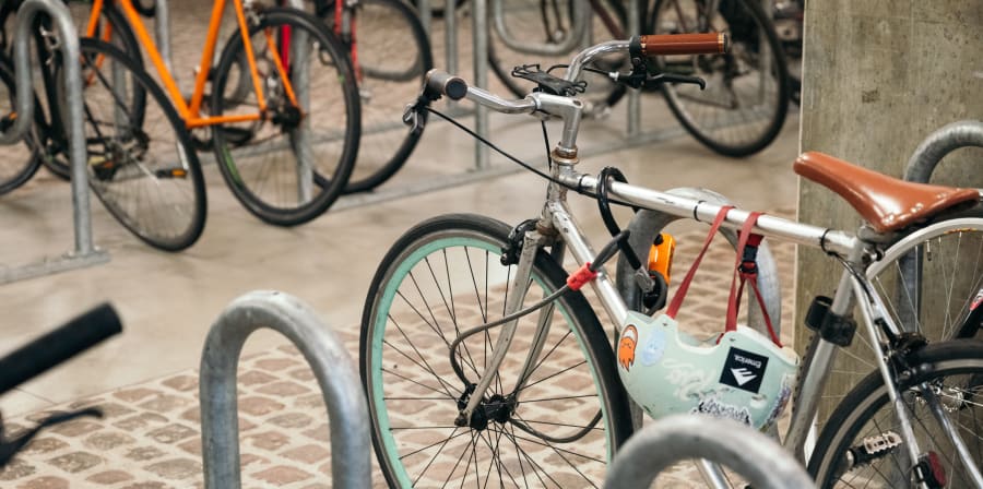 A silver bicycle with a brown seat is locked to a rack in an indoor area. A mint-green helmet hangs on the bike. Other bikes are visible in the background.