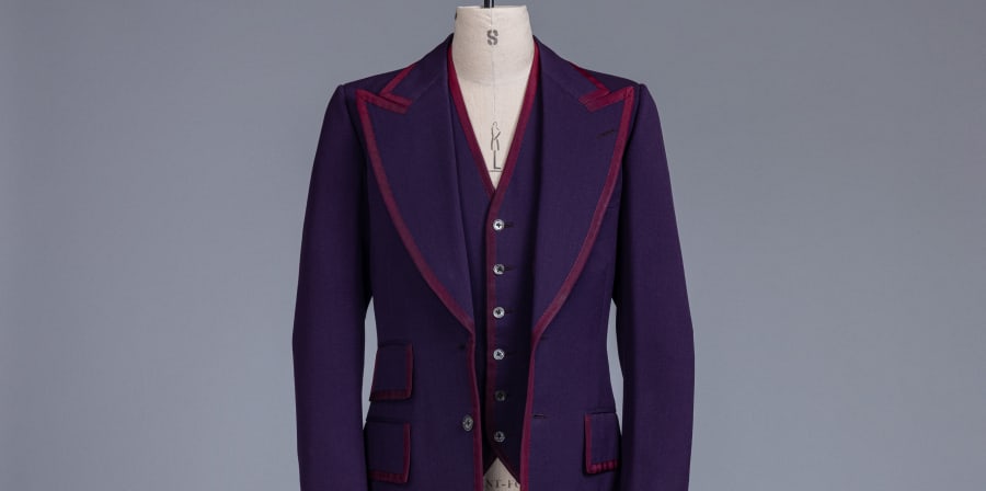 A two-piece suit jacket and waistcoat are mounted on a cream mannequin bust, against a white backdrop. The suit is a deep purple colour with a mauve piping around the edges, and exaggerated lapel.