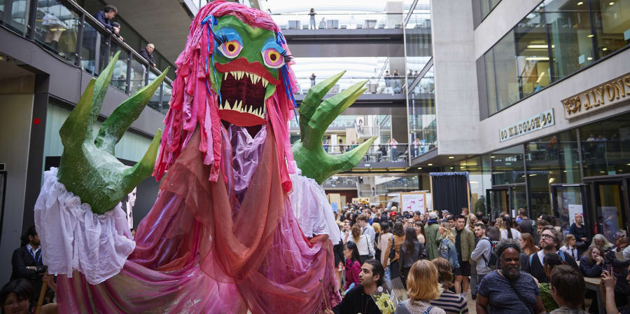 A huge pink puppet with green hands amid a crowd of visitors to the Street at Central Saint Martins