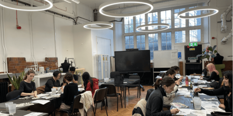 A group of people sit at several tables in a large, bright studio workspace