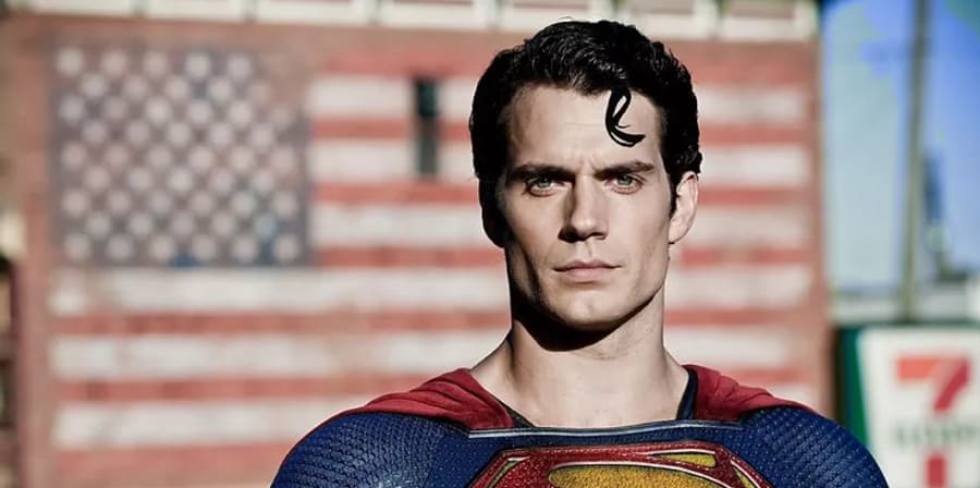 Henry Cavill as Superman in the film 'Man of Steel', standing in the Superman outfit in front of an American flag outside with a blue sky.