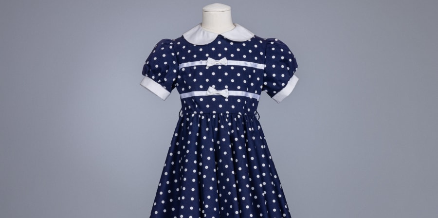 A blue and white polka dot child's dress with two white bows and puff sleeves and a peter pan collar on a cream mannequin bust.