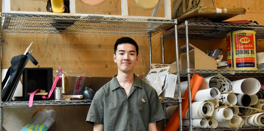 An image of student designer Arthur Yu. He is standing in front of a shelf hosting various print making materials. 