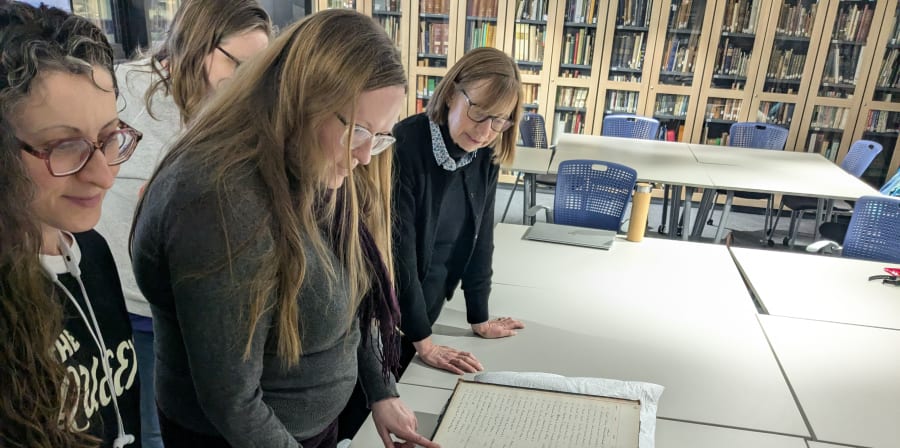 1 woman with long hair standing over a large historic register with other women looking at it