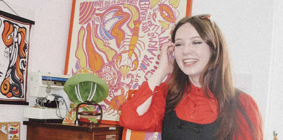 Smiling woman in a red blouse and black pinafore standing in a colourful, art-filled studio.