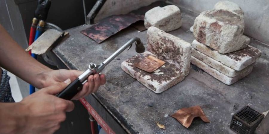 A pair of hands using a blowtorch on a small square piece of metal 