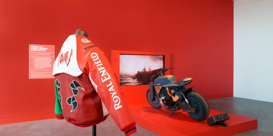 Exhibition space featuring a Royal Enfield motorcycle and a red and white racing jacket on a mannequin, both displayed against a vivid red wall. The motorcycle has a custom blue, orange, and black design, while the jacket prominently displays 