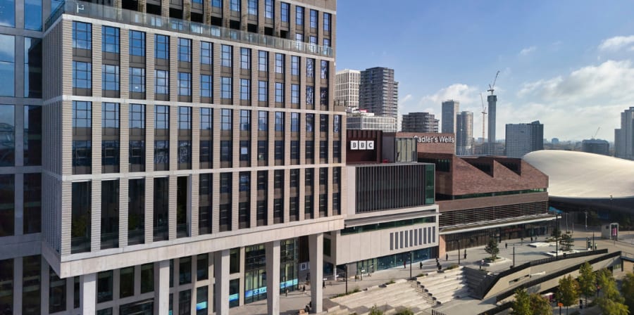 Modern urban landscape featuring the University of the Arts London (UAL) campus building in Stratford, with prominent signage from the BBC and Sadler’s Wells Theatre. The tall UAL structure is clad in glass and concrete with large grid-style windows, while adjacent buildings include the BBC studio with black and white logo panels and the angular brick facade of Sadler’s Wells East theatre. The architectural setting includes a wide pedestrian plaza, tiered steps, landscaping with trees, and surrounding high-rise apartment buildings. The distant skyline features construction cranes and the curved roof of the London Aquatics Centre.