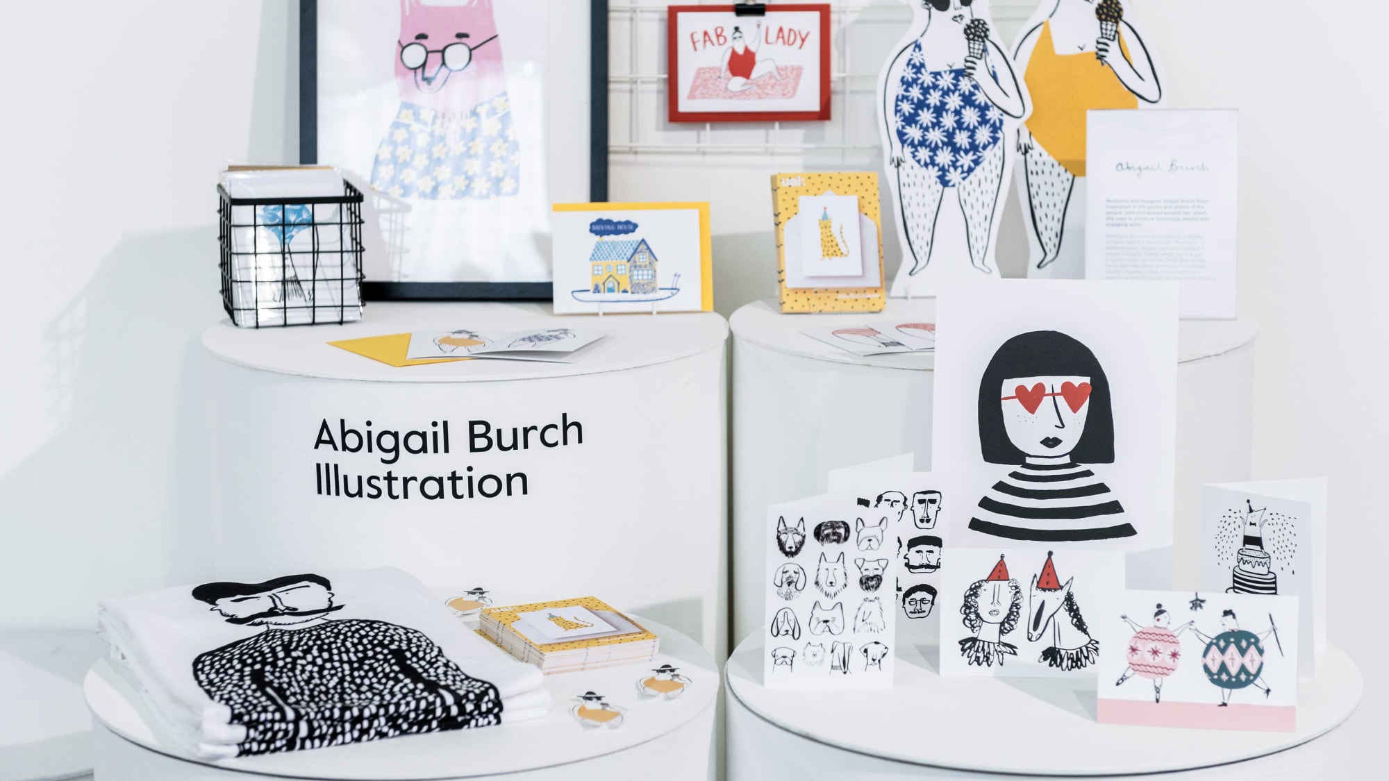 Display of Abigail Burch's illustrations at an exhibit. The setup includes framed artworks, greeting cards, and printed textiles featuring whimsical characters and patterns. The name 