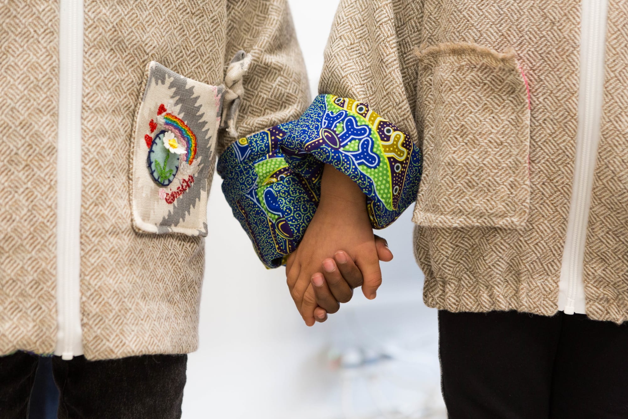Close up of two people holding hands. The focus is on the detailing of their jackets which features embroidery and brightly coloured batik prints