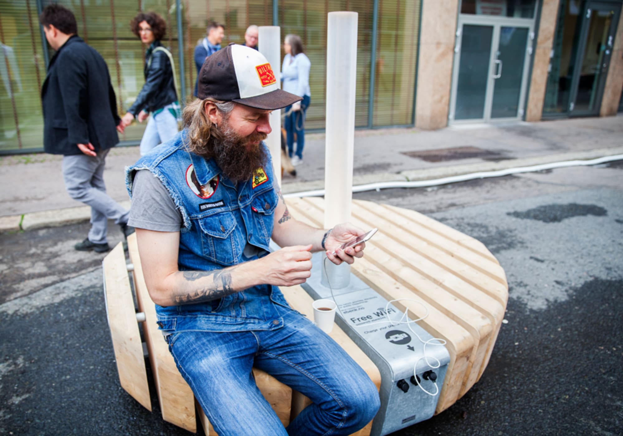 Man wearing baseball cap and jeans looks at his phone which is being recharged on the bench he's sitting on