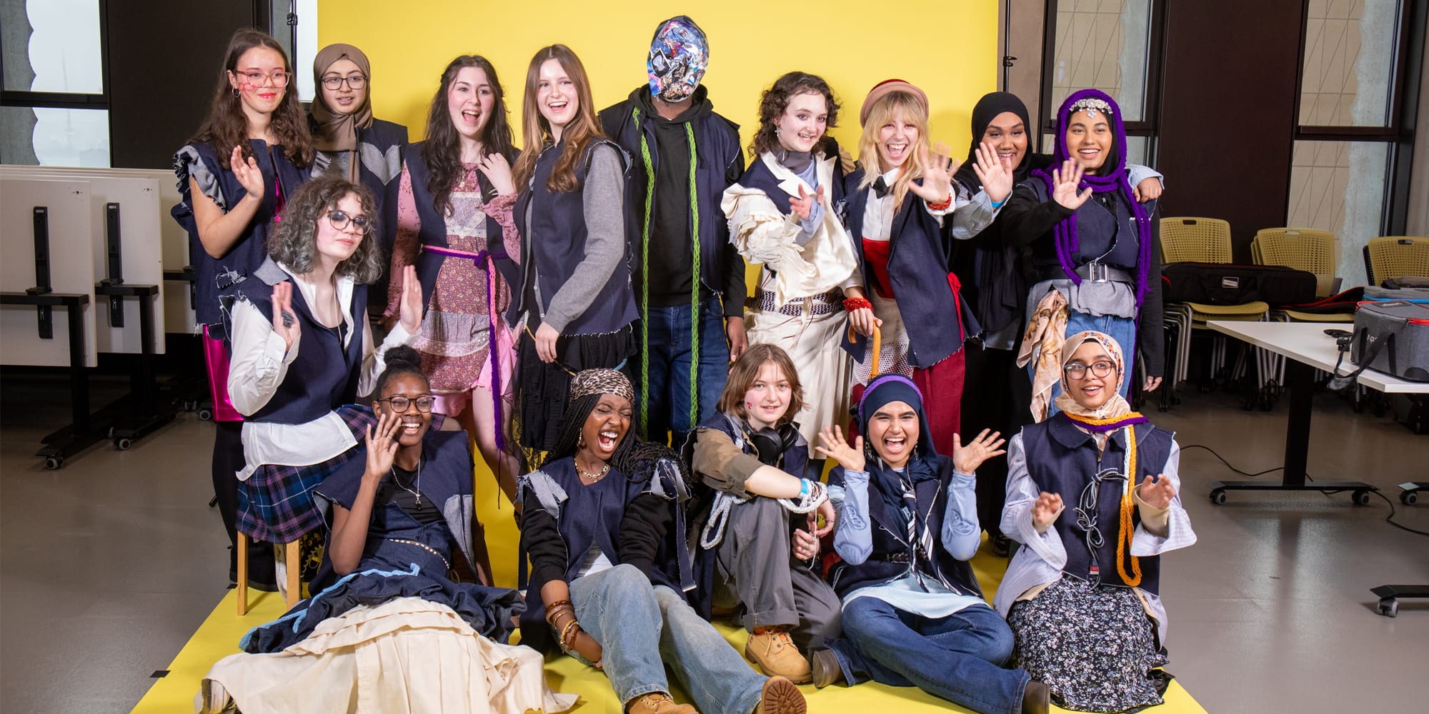 A group of students posing infront of yellow backdrop. They are wearing garments created during workshops.