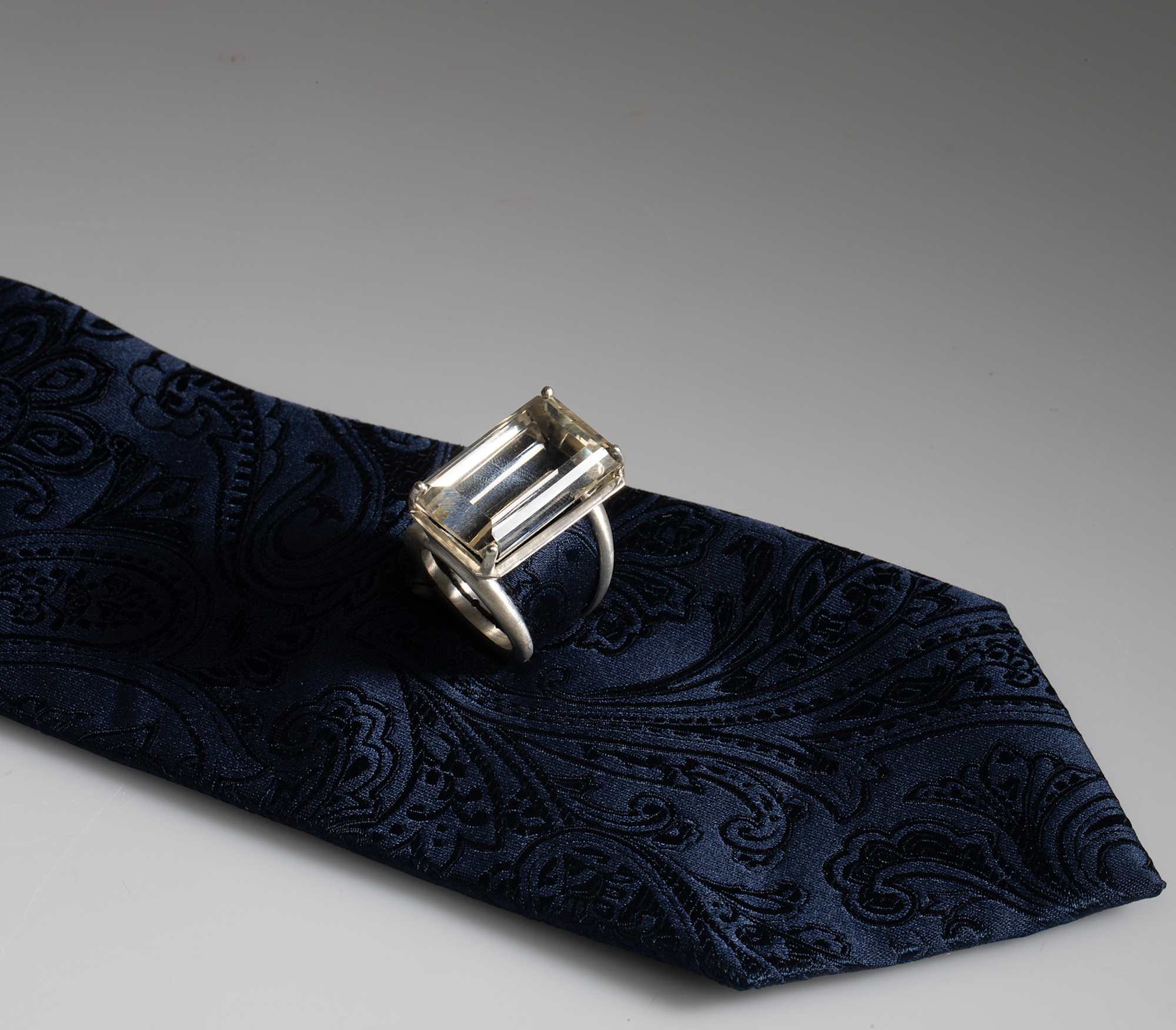 A silver ring with a baguette or rectangular style glass stone is placed on a textured navy tie