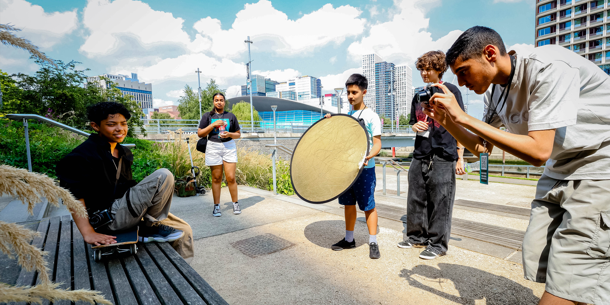 A group of students are standing outside working on a photoshoot together. Their subject is sat on the steps on a skateboard, in the background you can see the cityscape of Stratford