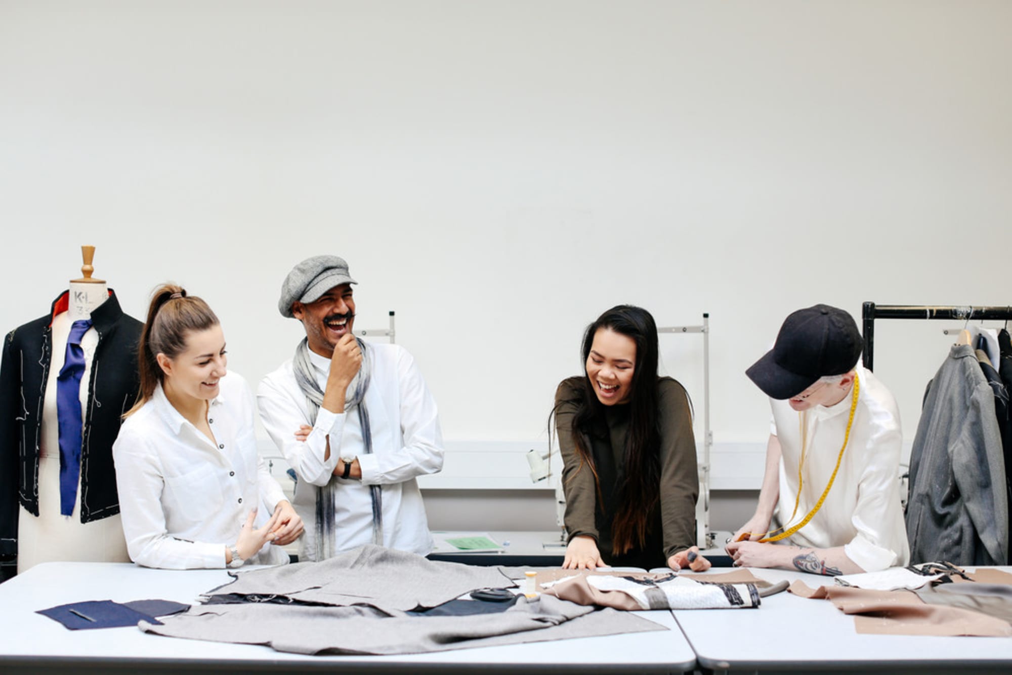 Four fashion students laugh and collaborate around a table covered in fabric and sewing tools in a bright studio.