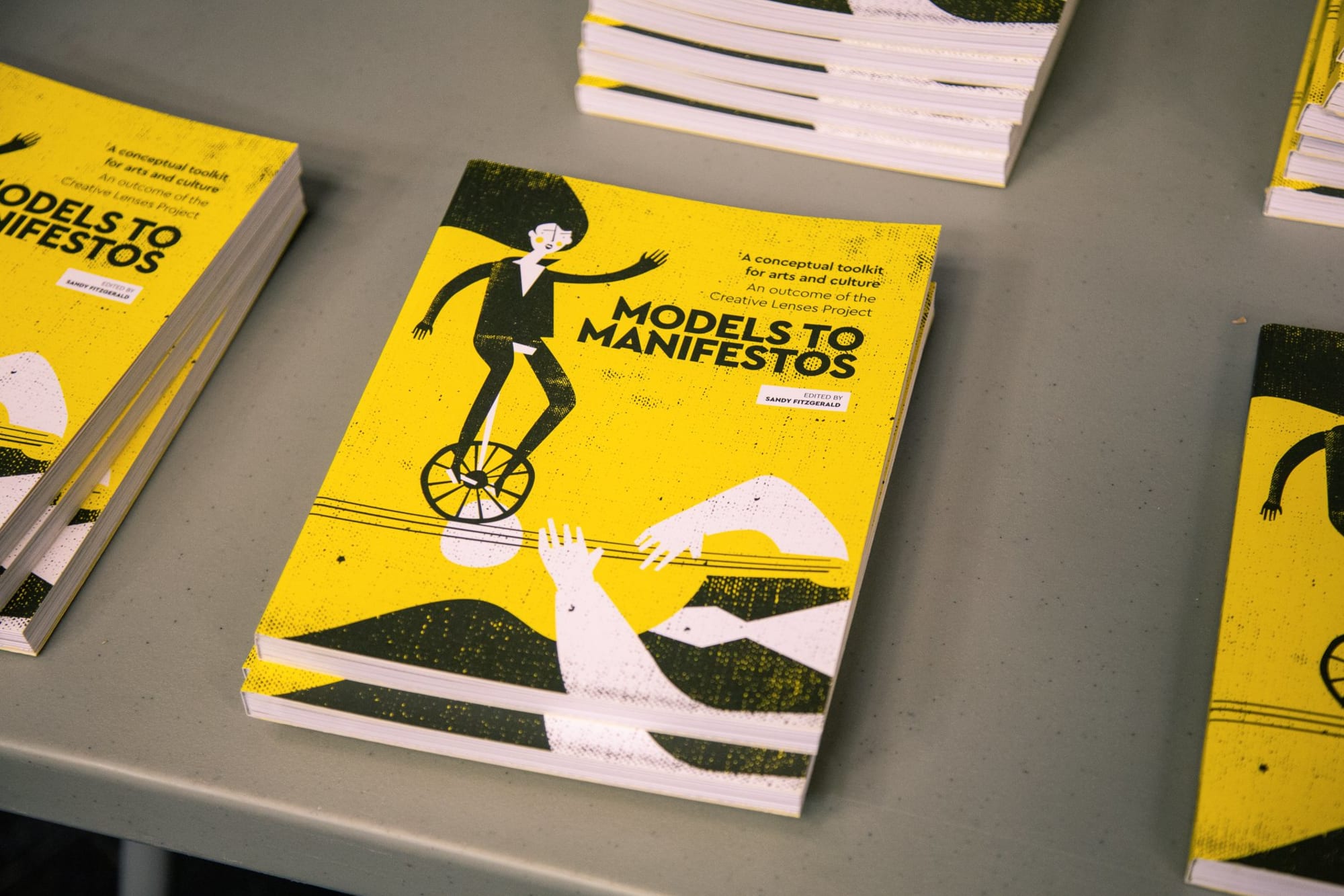 Small piles of the Models to Manifestos publication with a bright yellow graphic design on a table