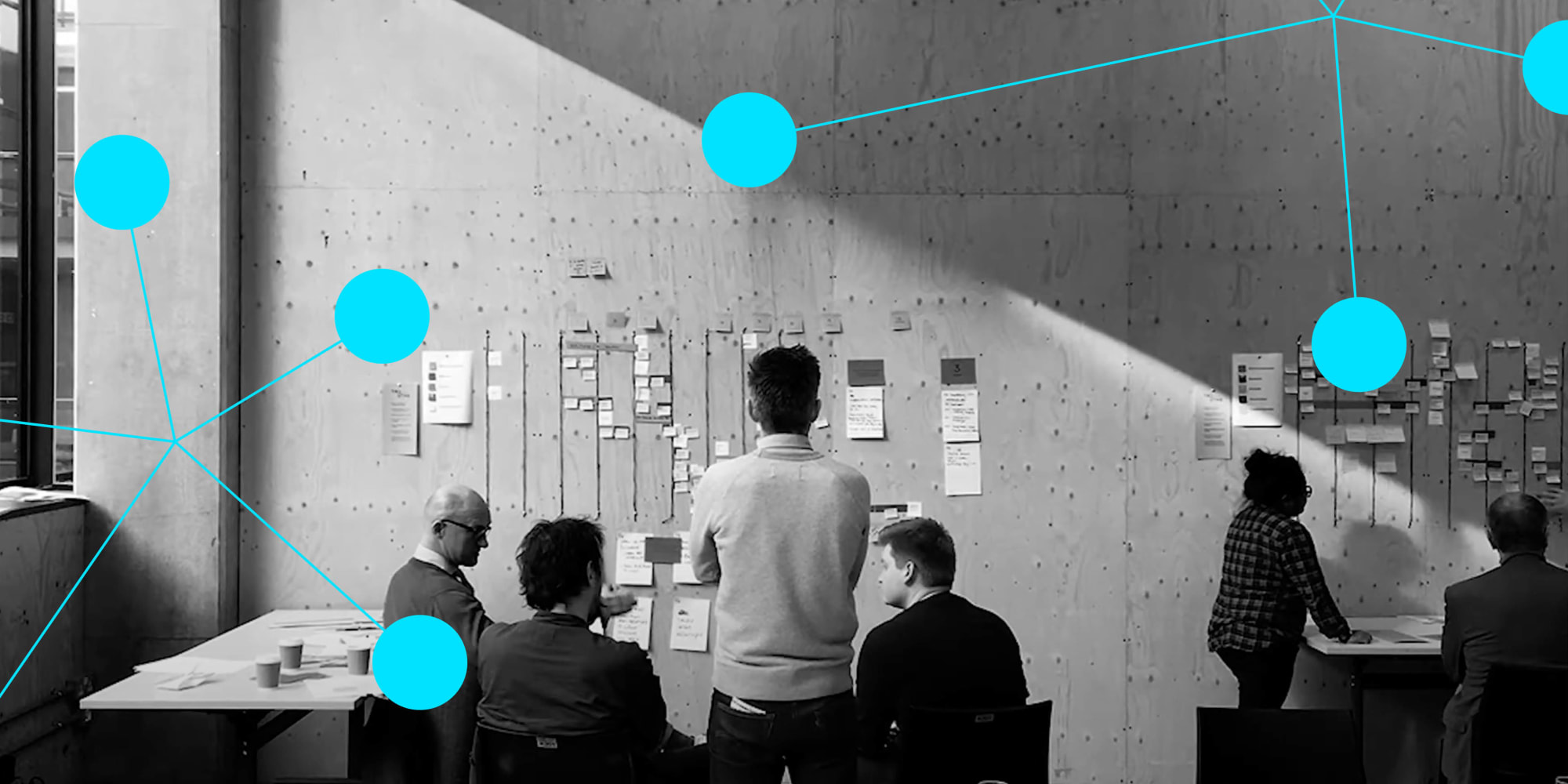 Black and white, a group of people in front of a wall that has post-it notes and plans on it. Light blue dots connected by thin blue lines overlay part of the image.