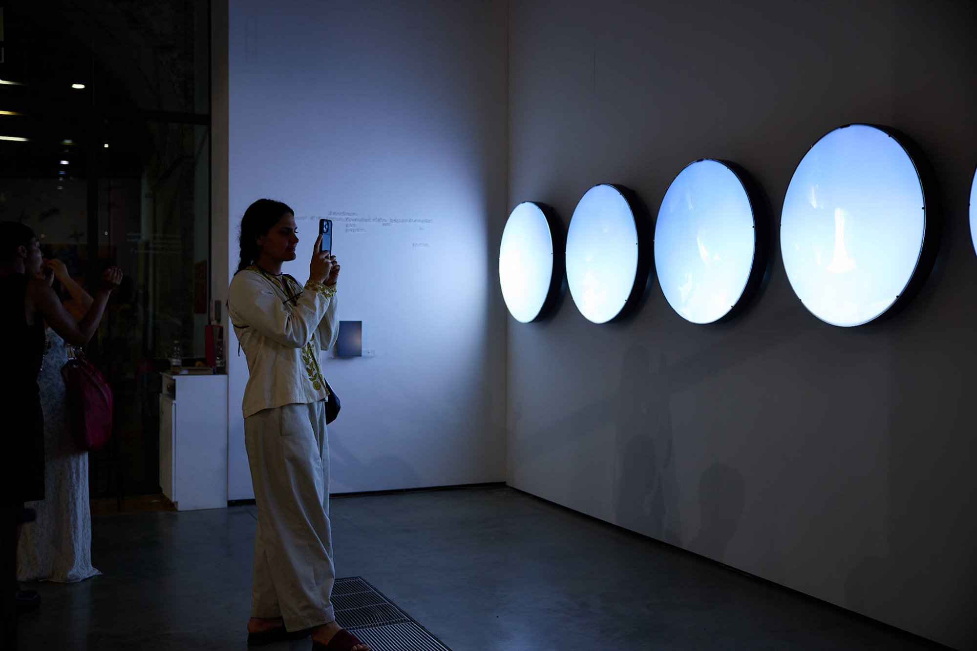 A person is standing in a dark space in silhouette taking a photo of a wall which is lit via an installation of a row of large round light boxes which have forms visible on each orb and are emitting a blueish light