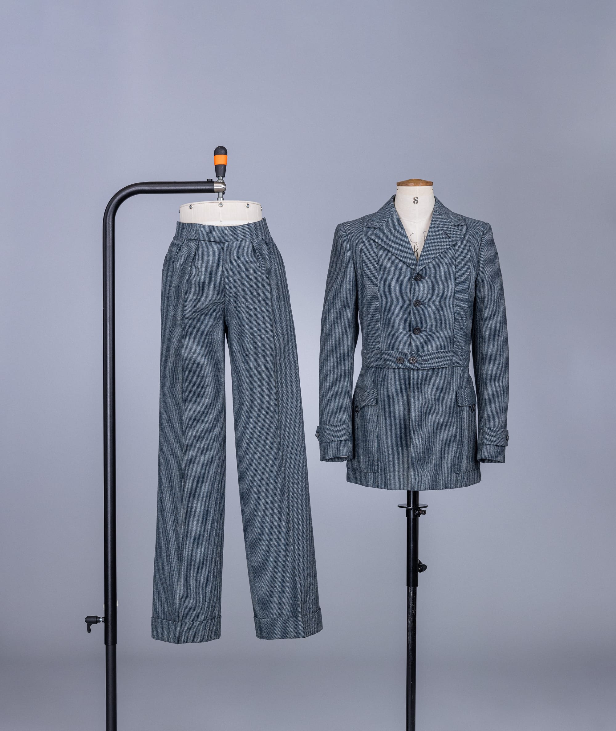 A grey two piece suit, on separate cream bodice and legs mannequins, supported by black metal stands, on a white backdrop