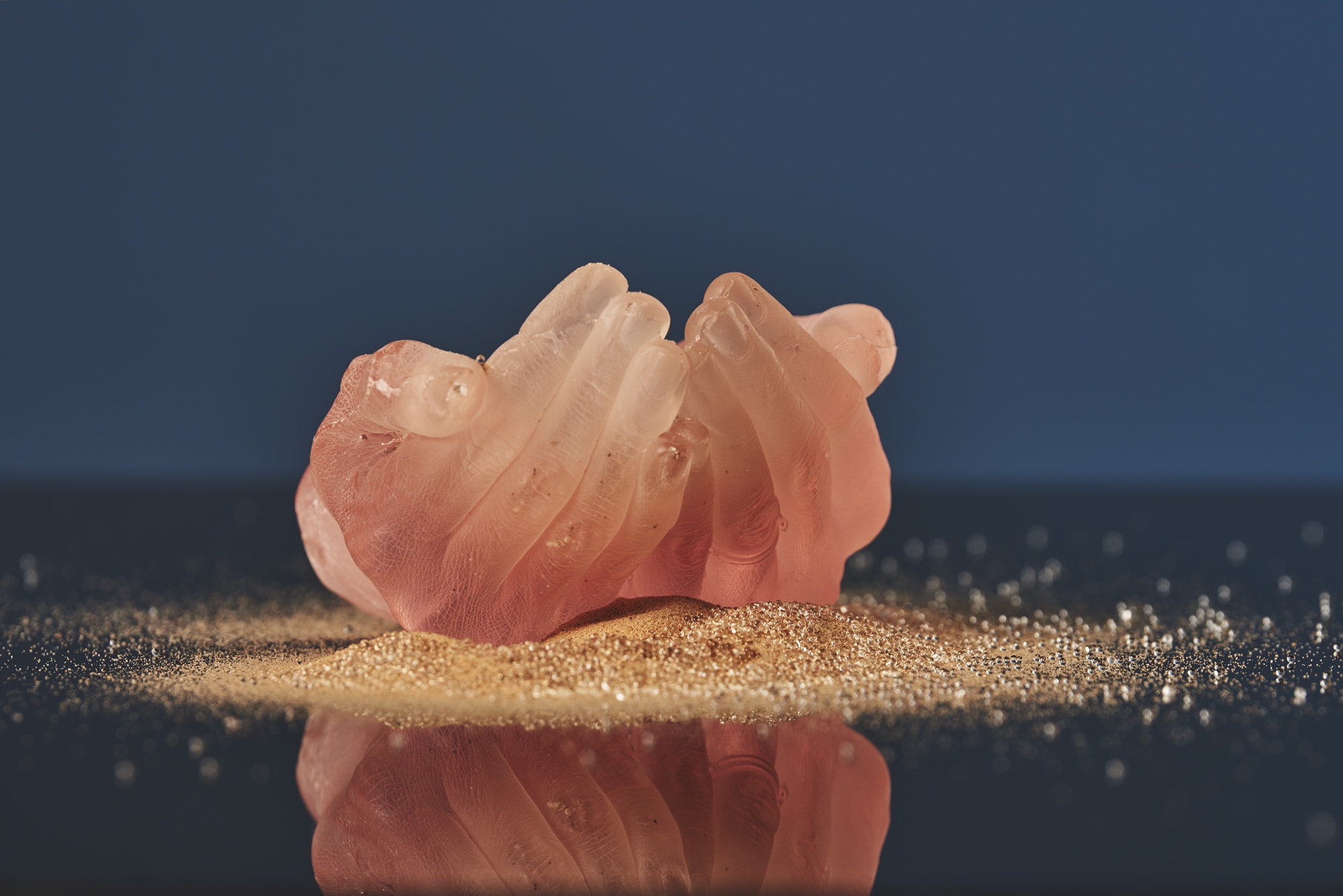 Pink translucent sculpture of cupped hands on a reflective surface with sand, against a dark blue background.