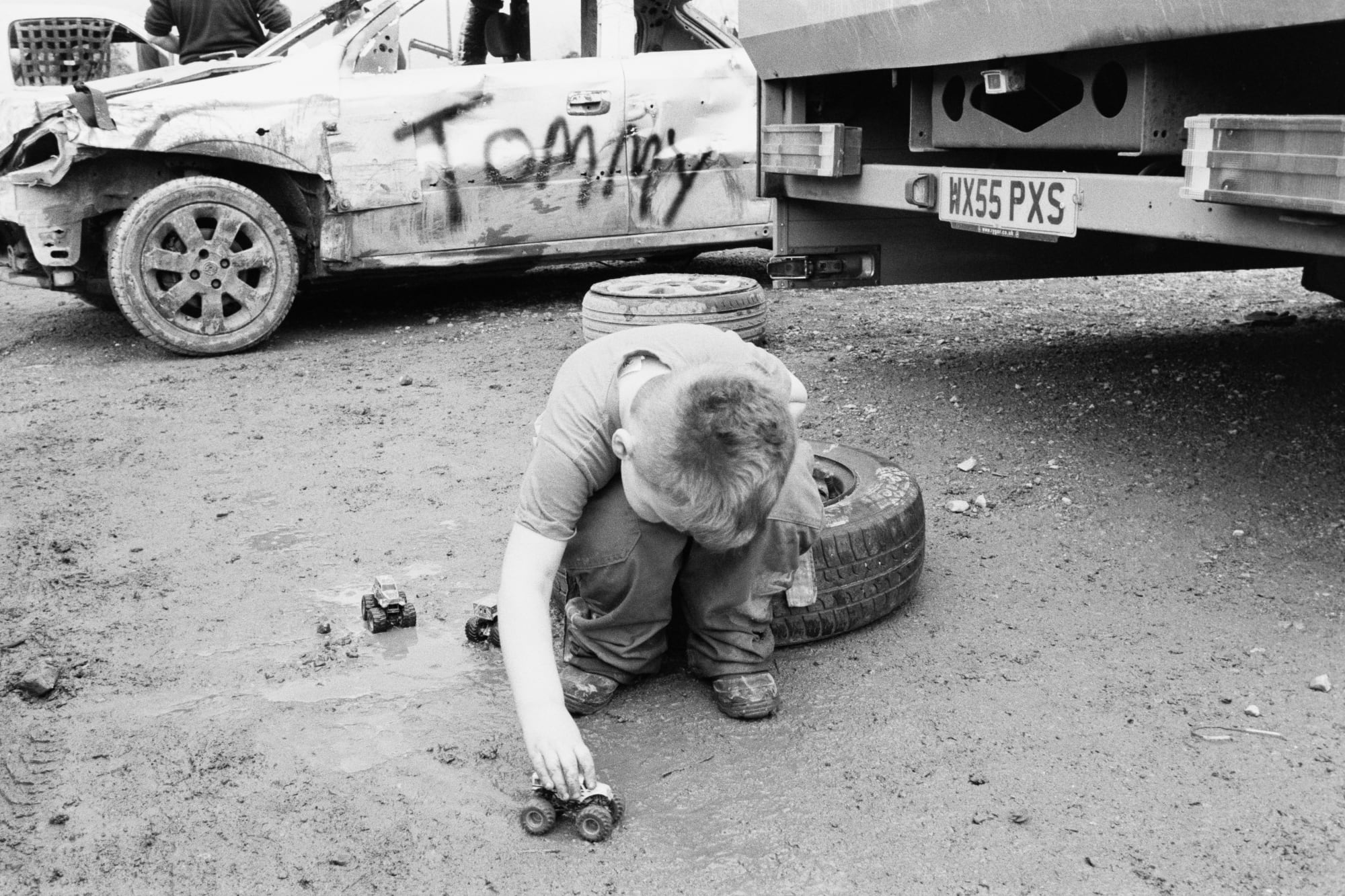 A greyscale photograph of a small child crouched on the ground playing with a small truck. The child is seated on a car tyre and visible in the background are the sides and cars and the registration plate of a truck. The car has the word TOMMY in caps graffitied on the side.