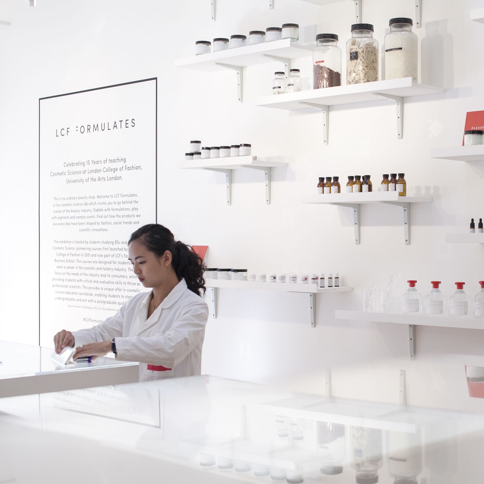 Woman in white lab coat with shelves of cosmetics behind her. LCF Formulates Cosmetic Science exhibition, Fashion Business School Summit, Kingly Court. Copyright holder: Emmi Hyyppa
