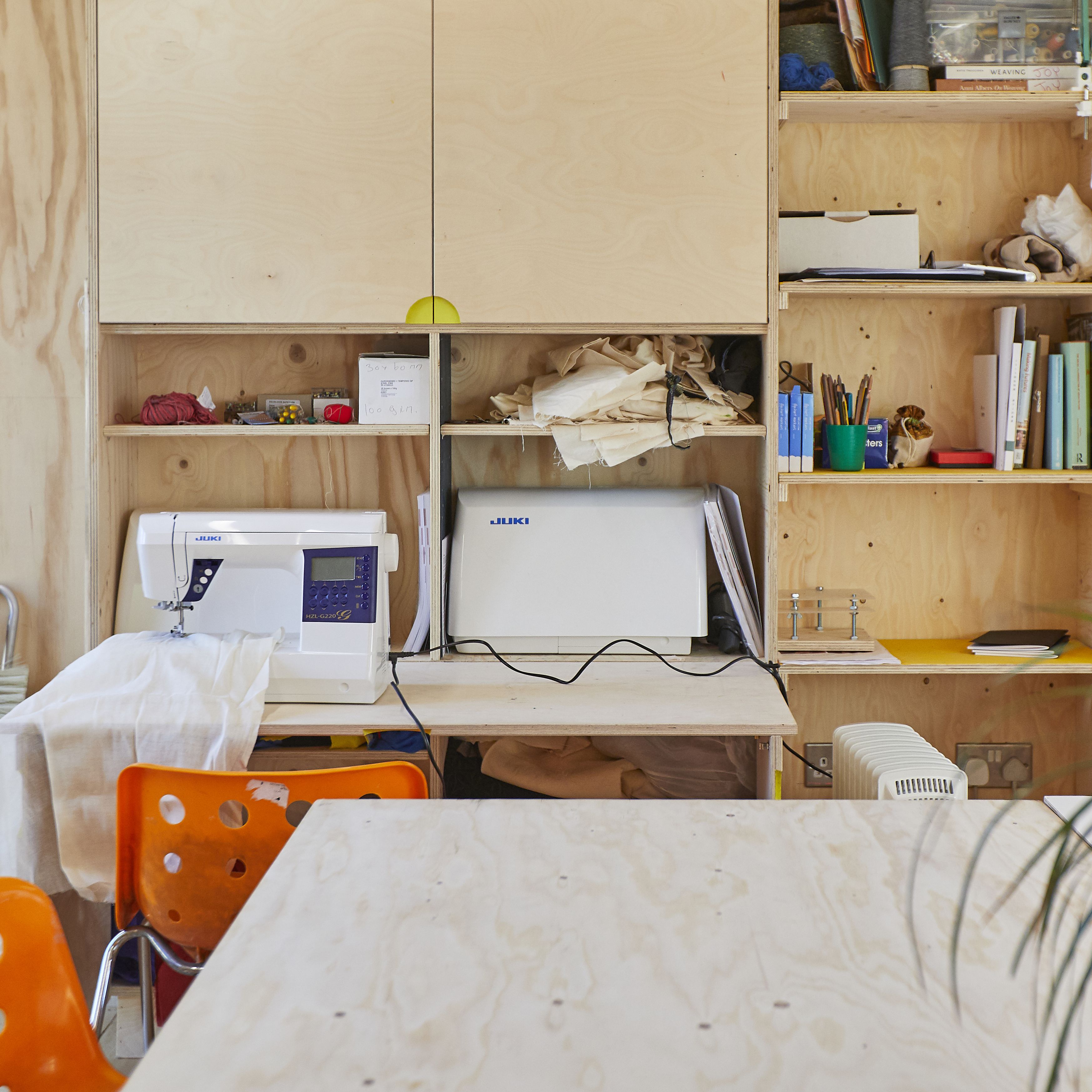 A informal but well organised workshop with plywood walls and cupboards, orange chairs, a sewing machine and a tabletop in the foreground