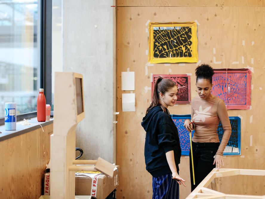 2 female students preparing for an exhibition, one holds a measuring tape, and in the background there are plywood walls and surfaces. There are 2 posters on one of the plywood walls