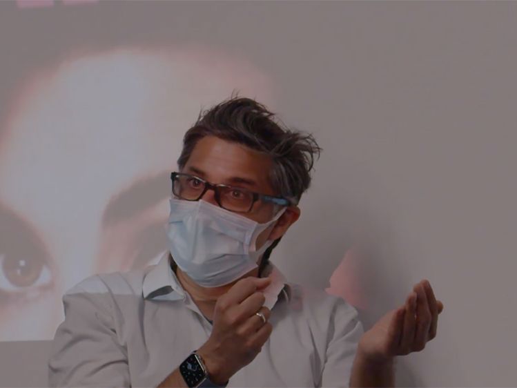 A director wearing a face mask sits in front of a projected screen.