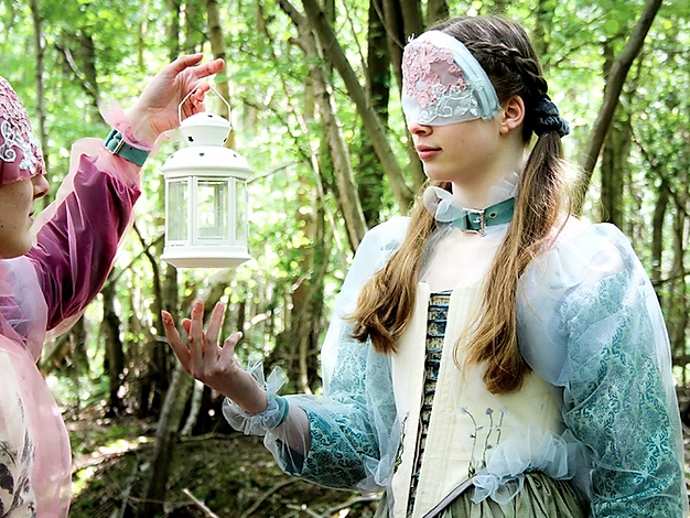 Two masked models interact with eachother while in a forest looking away from the camera wearing garments inspired by the pre-Raphaelite art movement