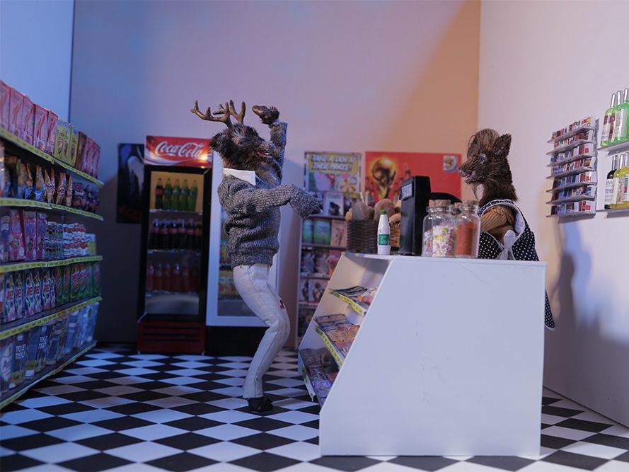 A male model of a deer, taken as a still from a stop motion animated film, waves to another deer in a shop scene.