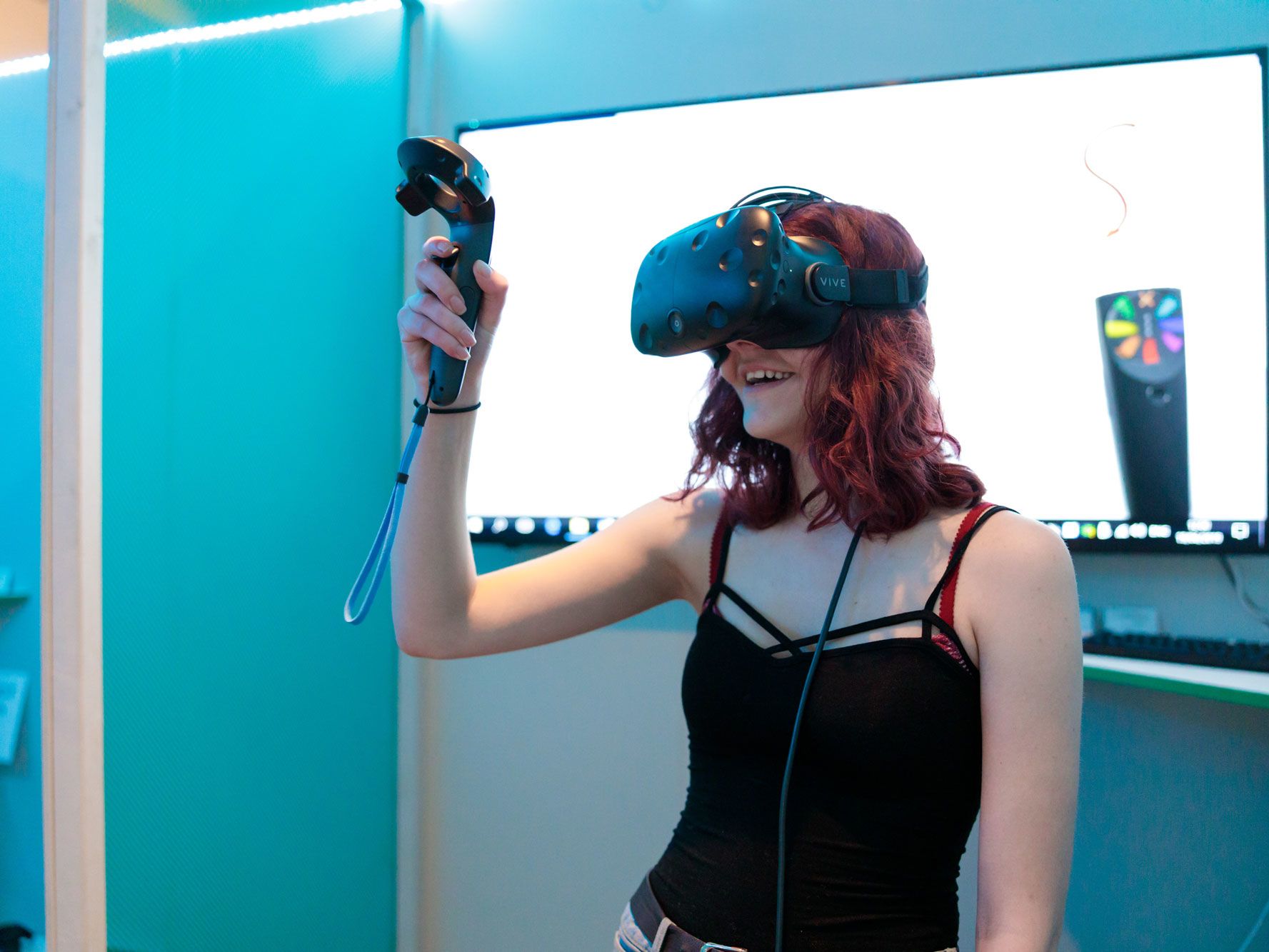 A girl in a black vest stop stands against a blue background wearing a VR headset and holding the controller in her hand, looking upwards.