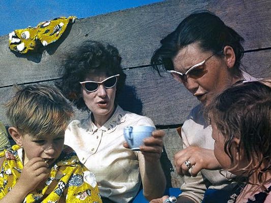 A family, wearing sunglasses, sit on a beach and drink tea.