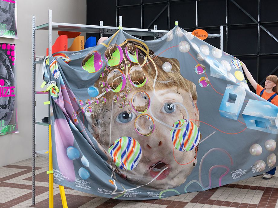 An artist stands beside a bedsheet-sized artwork depicting a face surrounded by coloured bubbles.