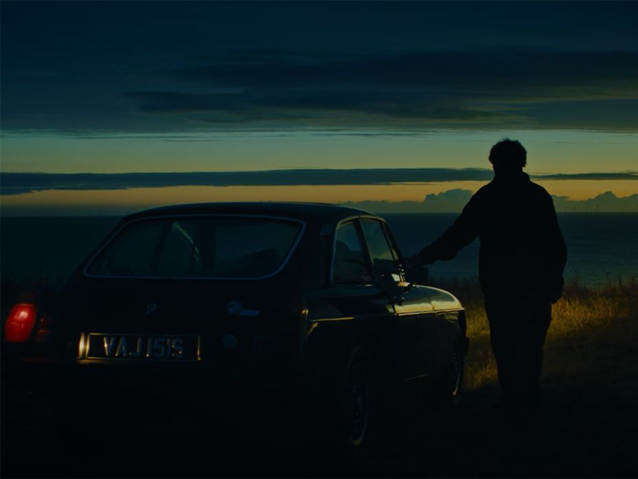Still of a man standing beside a car at dawn.