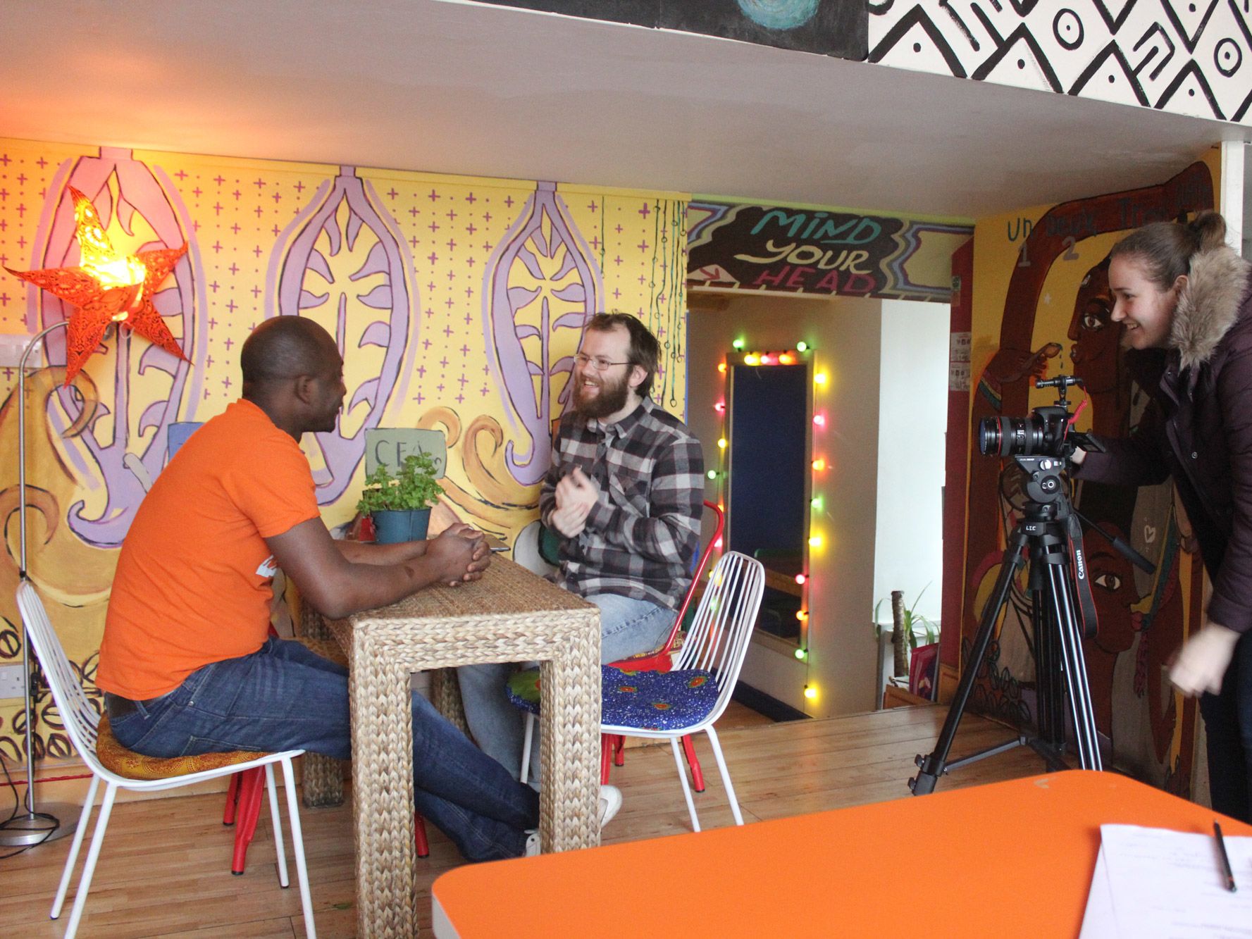 Two men, one client and one student, sit on a table in a cafe while a camera-woman films
