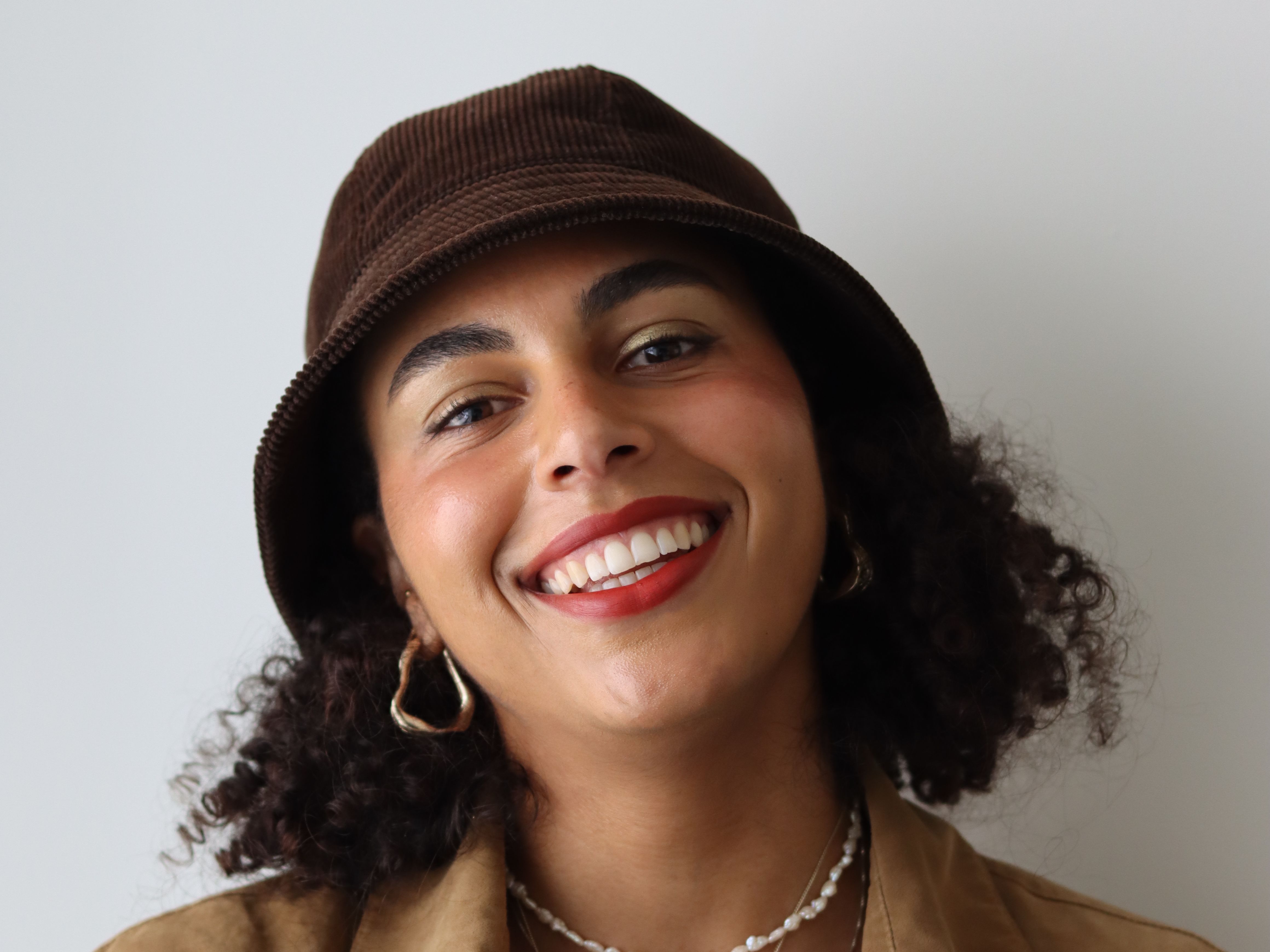 A person wearing a bucket hat and smiling with their chin tilted up. They are standing against a white wall.