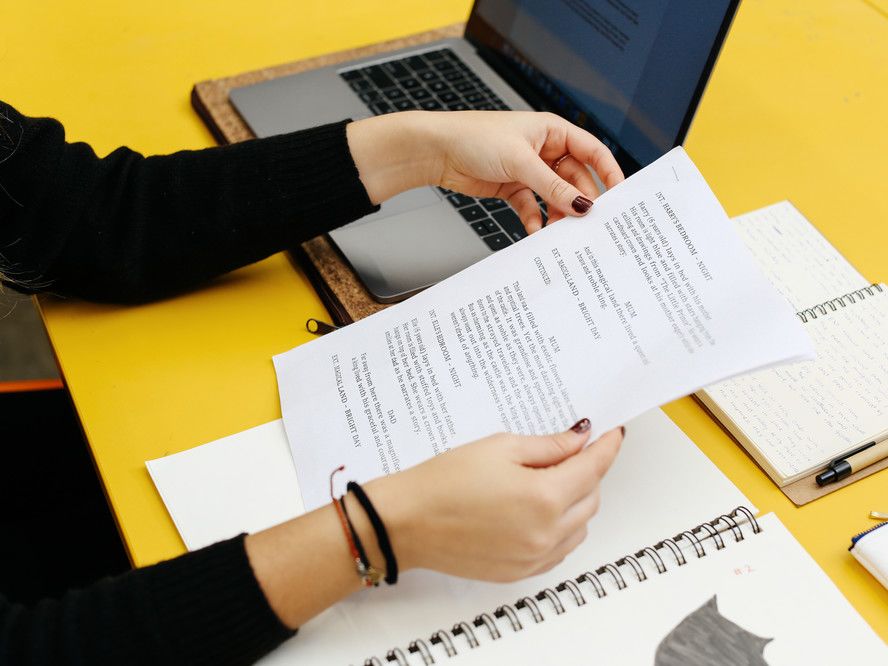 Close up of a woman's hands holding papers against a yellow desk with a laptop and notebooks
