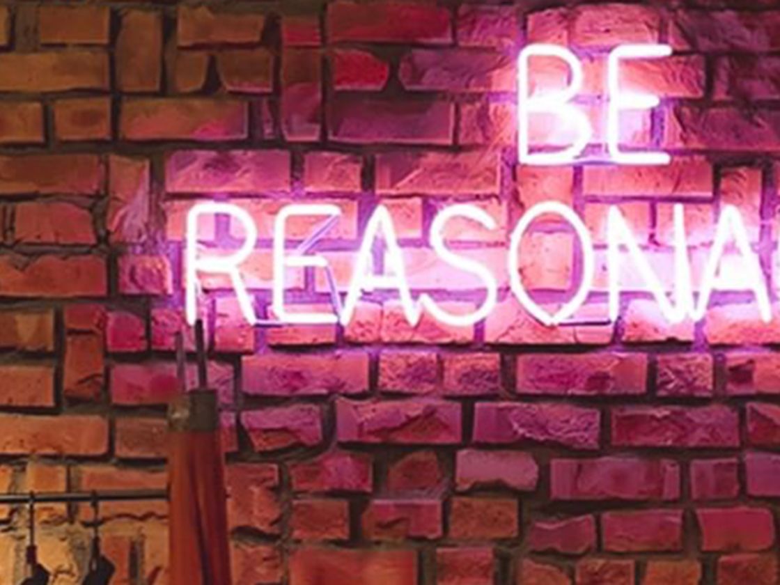 Be reasonable written in pink illuminous lights against a brick wall