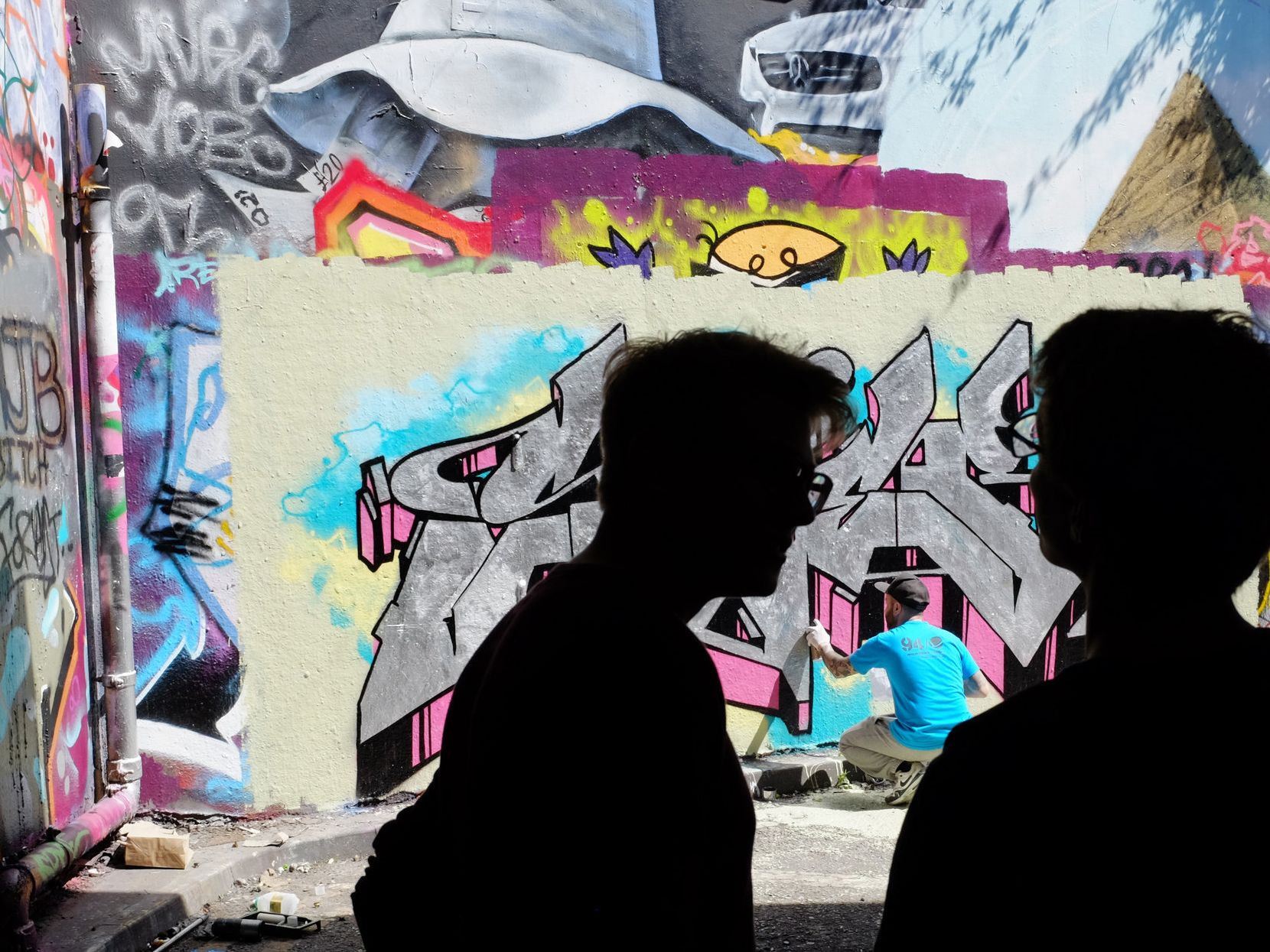 Two people watch a graffiti artist create work on an inner-city wall.