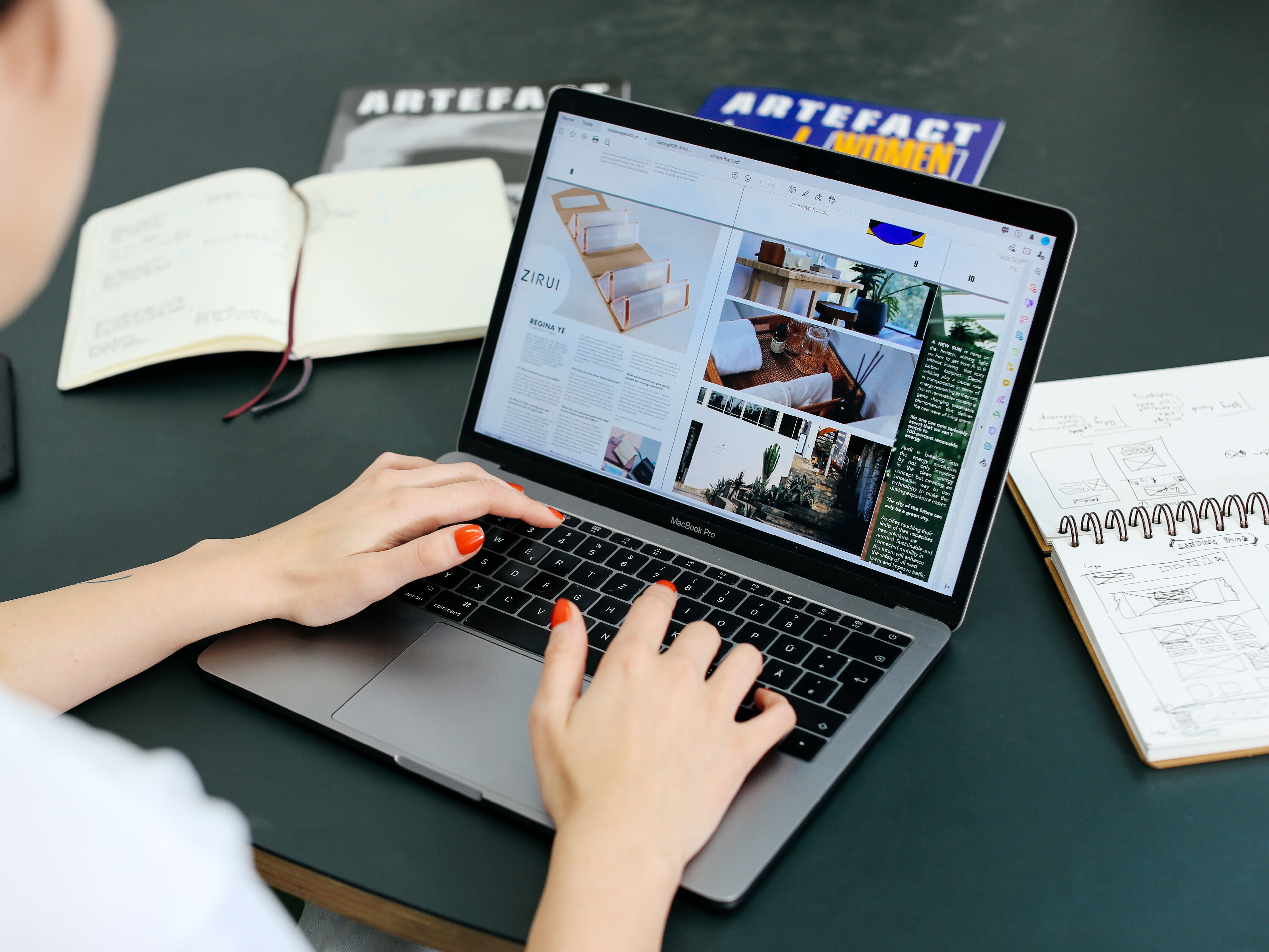 Person typing on laptops with 2 notebooks and an open magazine on the table.