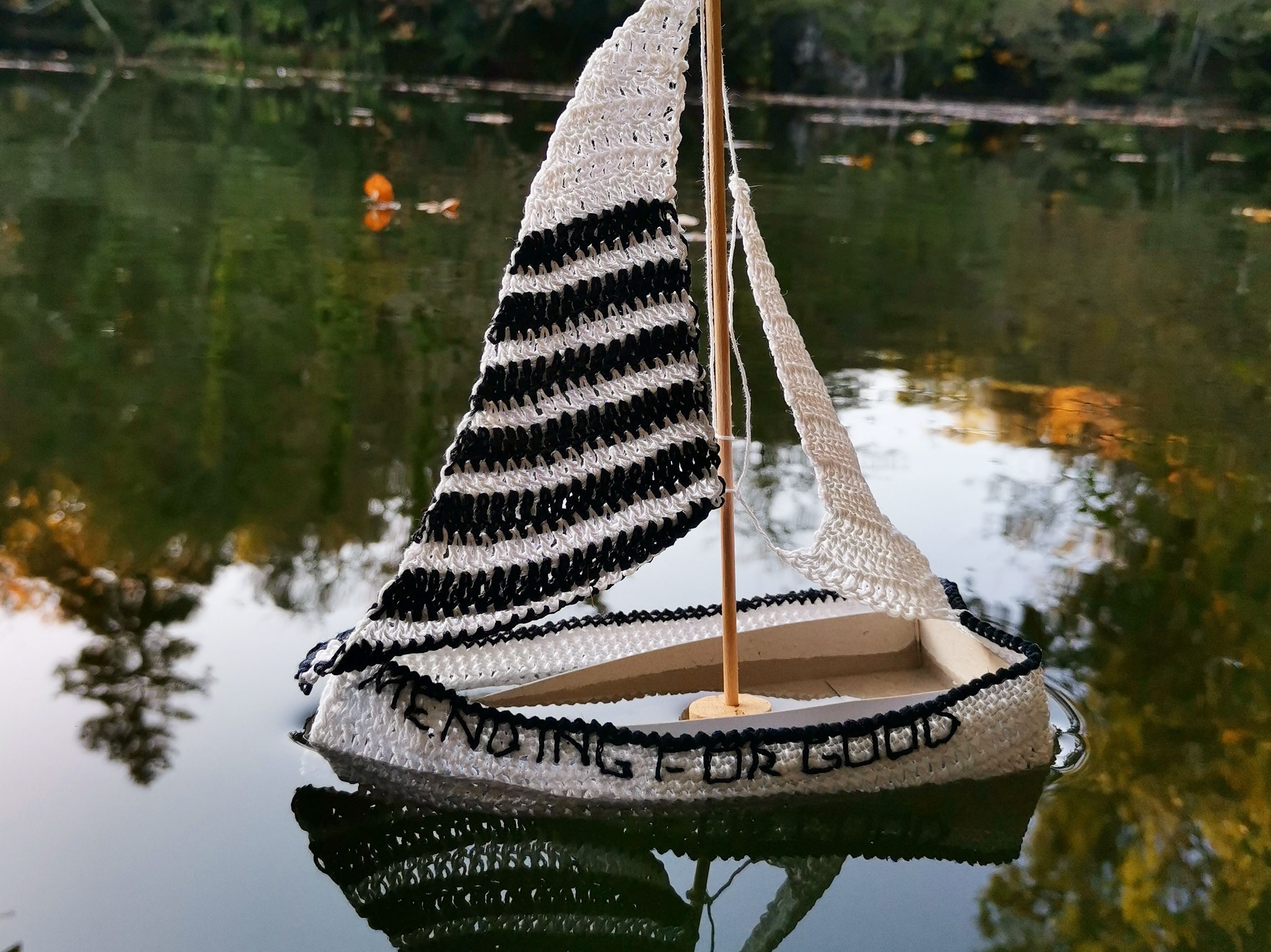 wooden model sized boat covered in a crochet monochrome design, with crocheted sails, floating on a body of water