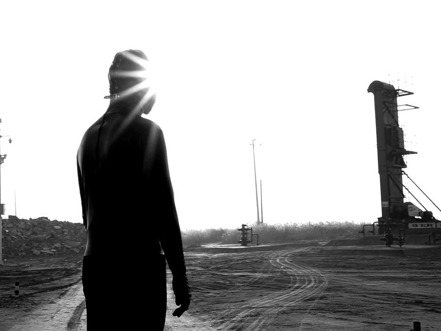 Person in a leather mask stands in front a industrial site in this black and white photo