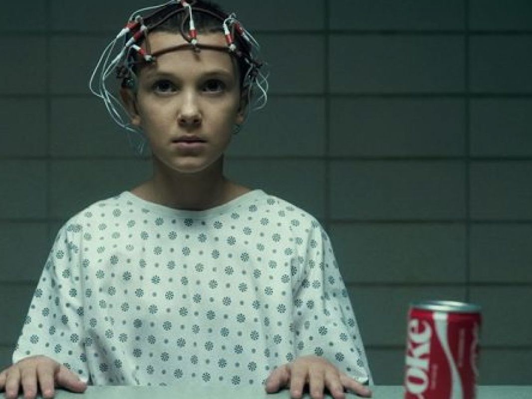 Still from the television programme Stranger Things, showing Eleven sat with an electrode helmet by a table with a Coke can.