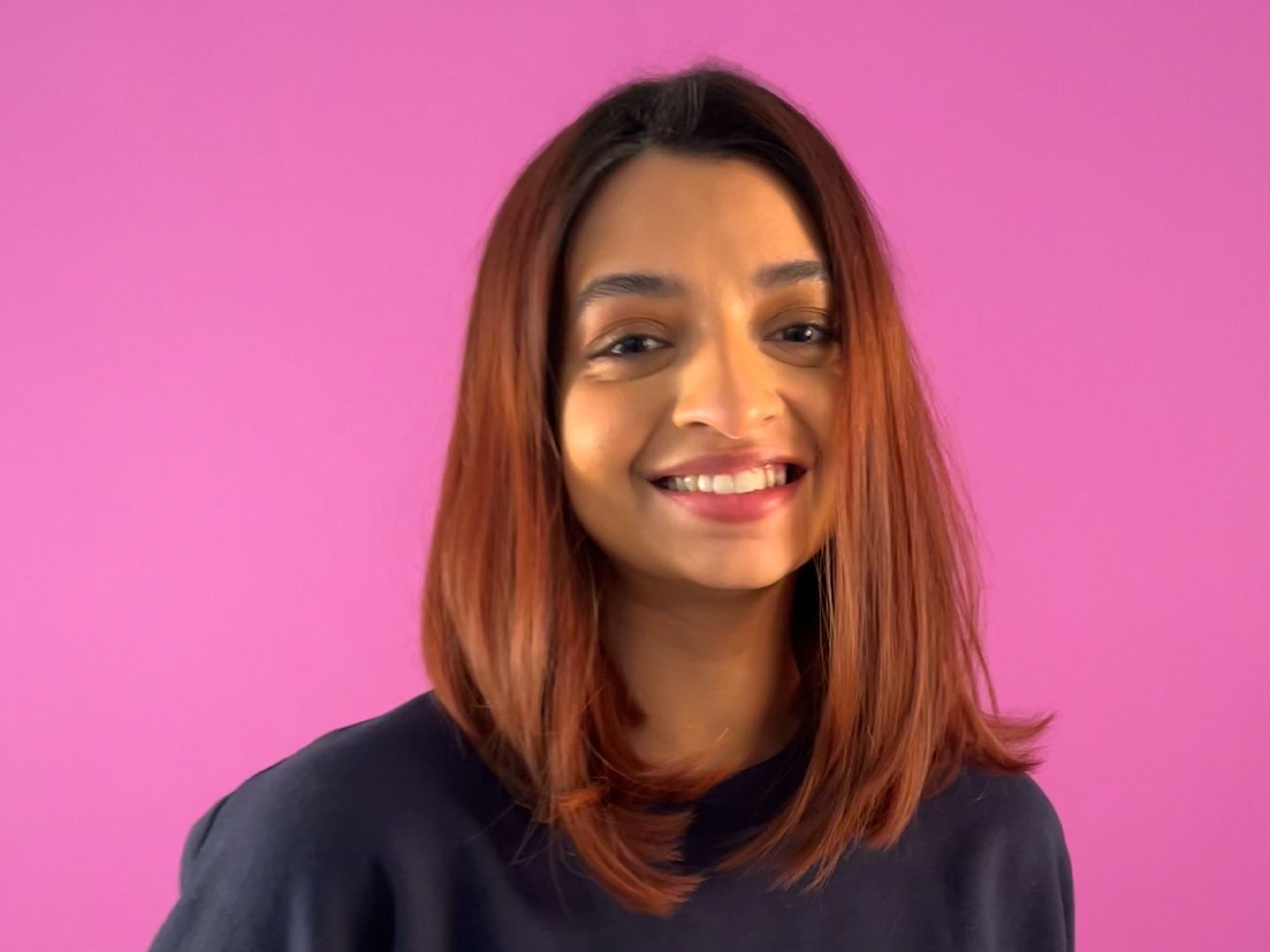 A young womanin a dark blue sweater smiles at the camera ,against a pink background
