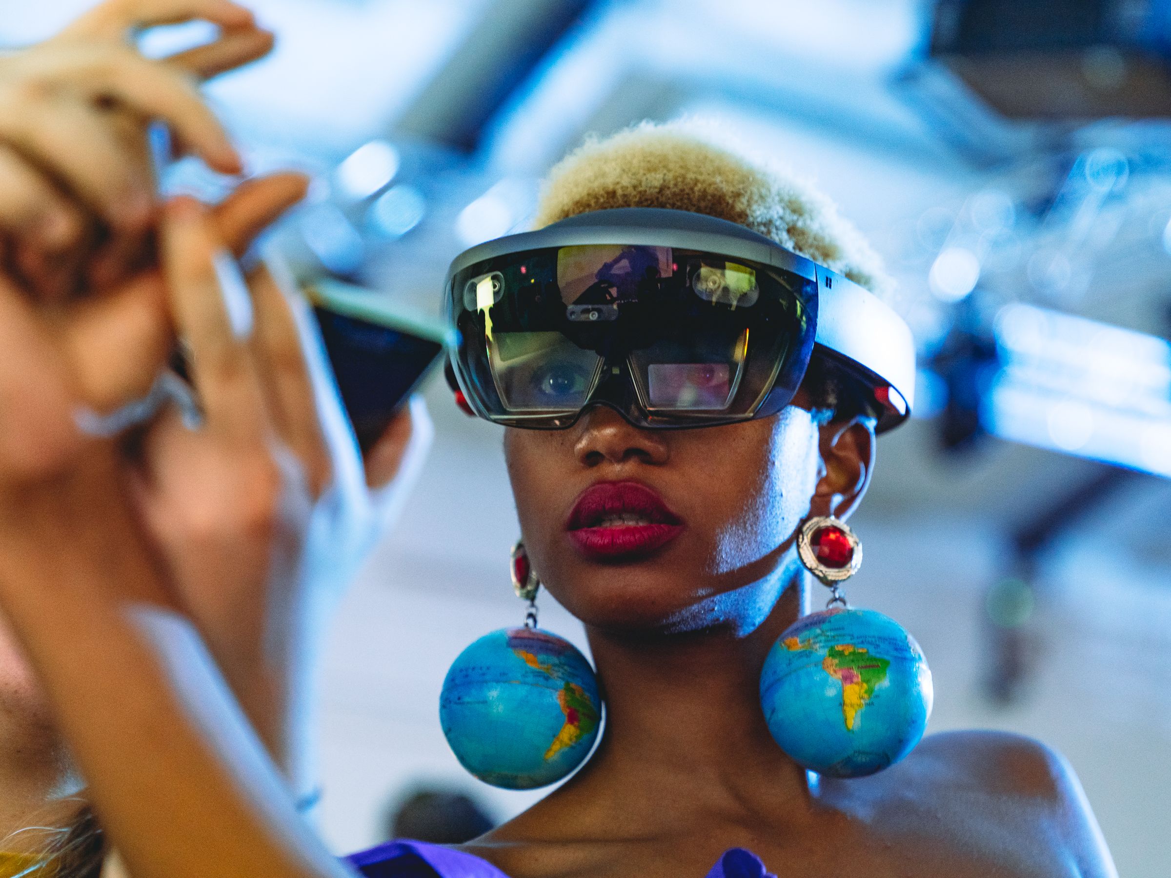 A woman with blonde hair wears virtual reality goggles. Her earrings are large globes each with a map of the world. 