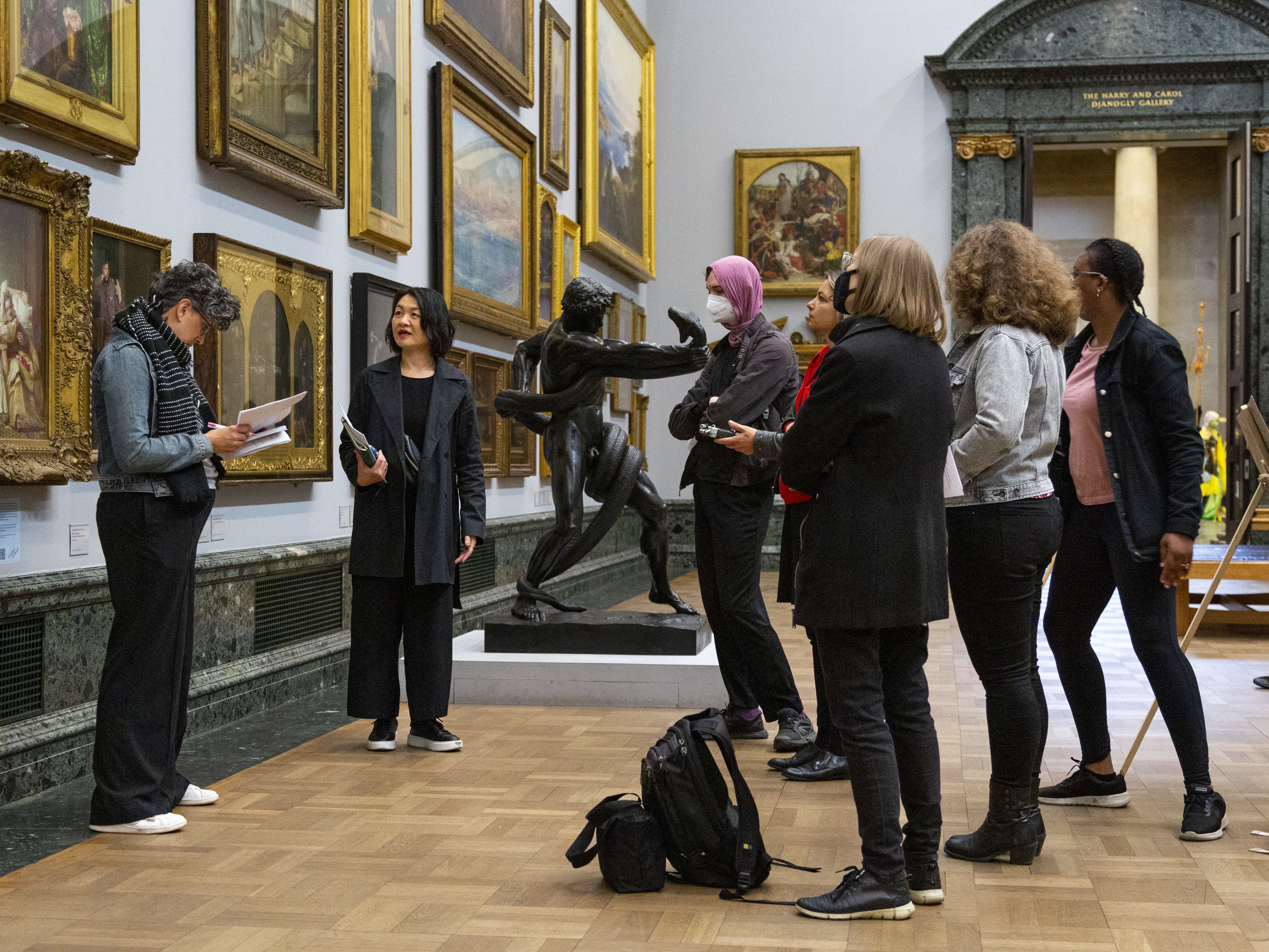 A group of people in a museum looking at ornately framed artworks on the wall. There is a parquet floor and a doorway in the background with pillars 