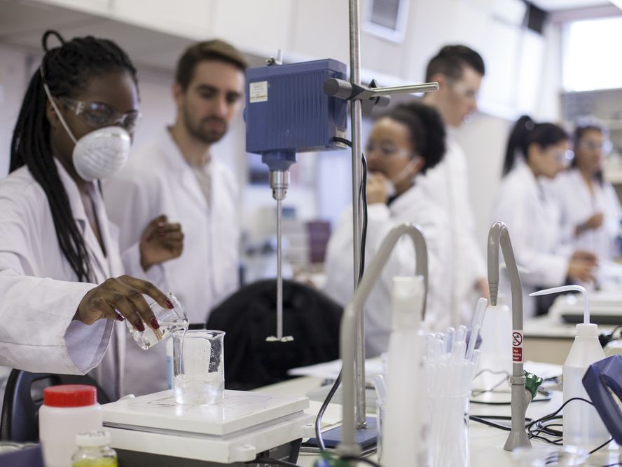 Cosmetic Science students from London College of Fashion in a lab.In the foreground a person is pouring clear liquid into a glass beaker. 