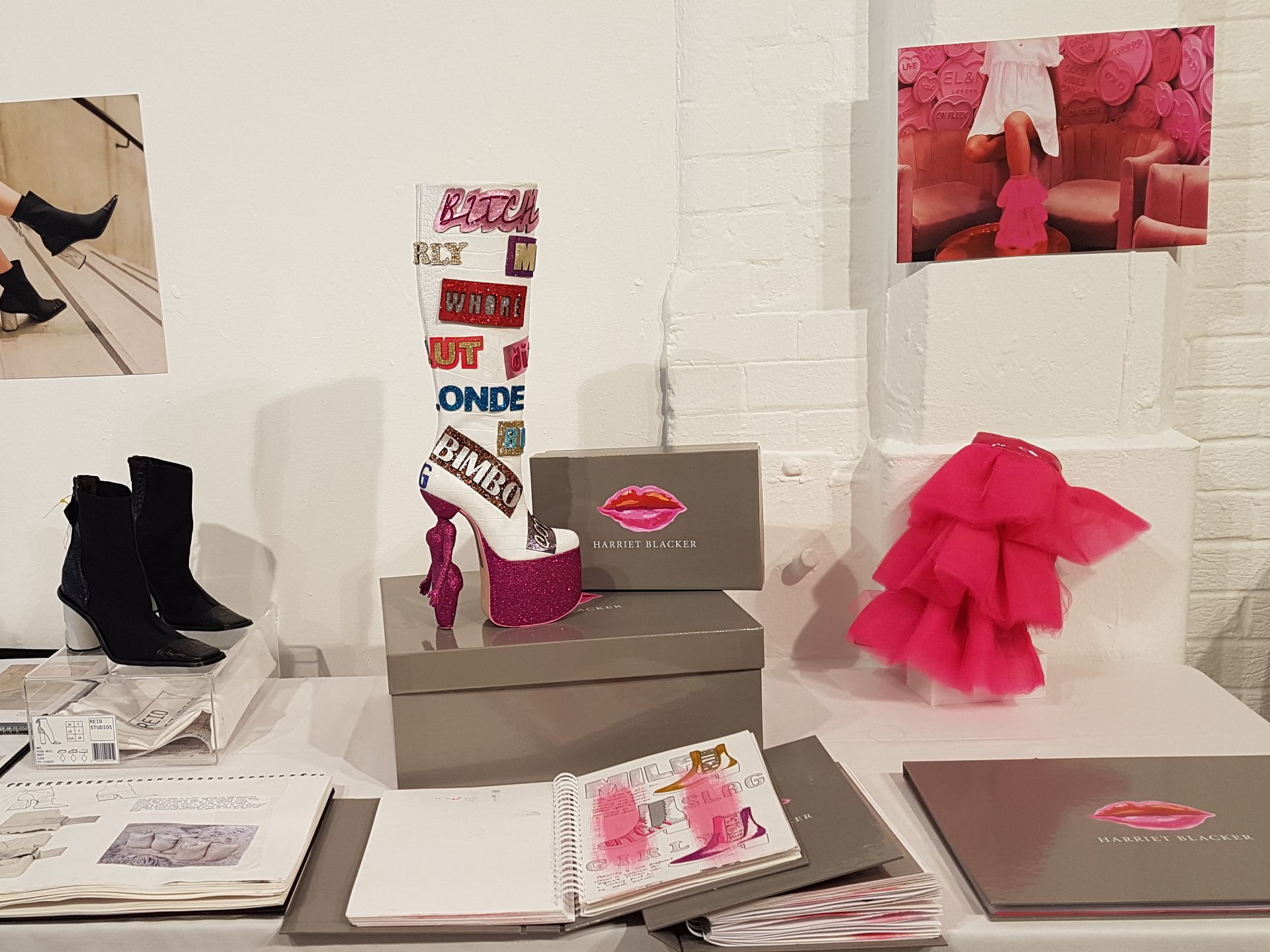 A black ankle boot with a concrete white heel stands next to a high heeled knee high boot in pink glitter which has badges on it with words like 'bitch' and 'bimbo.'